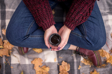 Young woman holding a cup of coffee, sitting in the park on a plaid. Autumn season.