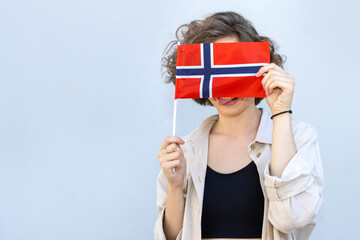Cheerful young girl showing her tongue hiding behind the flag of Norway, standing against the...