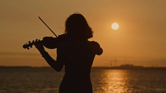 Silhouette of woman musician playing violin outside, standing in counter sunrise light, hair is fluttering in wind, harmony of nature and music art, Slow motion.