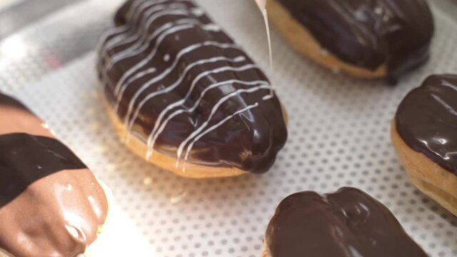 Close-up shot of a baker glazing the eclairs with white chocolate at the bakery. Delicious fresh sweet pastry. Production line at the confectionery factory. Making chocolate dessert. Food industry.