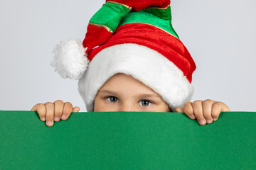 close-up portrait of mischievous child in Christmas gnome hat hiding behind black green poster with copy space in hands, isolated on white background. 