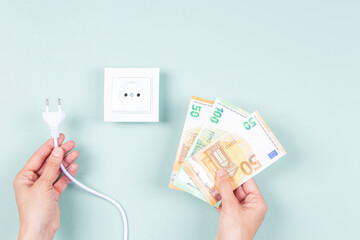 Fototapeta Electricity cost and expensive energy concept. Female hands holding electric power plug and Euro money banknotes near white electric socket on light green background obraz