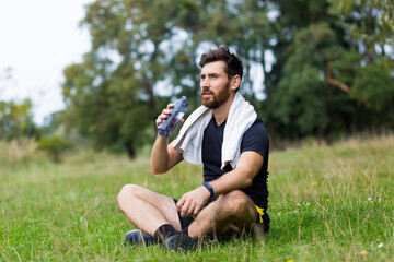 Exhausted after workout training or jogging athlete man sitting outdoors with a towel. Portrait sweaty fitness male in city park background forest and trees. Caucasian sportsman resting after running