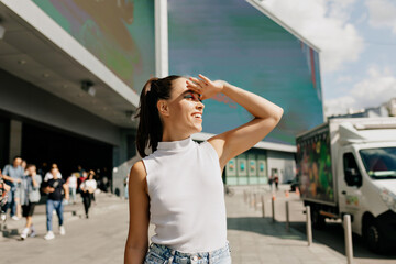 Trendy portrait of young stylish girl posing outdoors. Adorable brunette is looking aside and smiling on city background