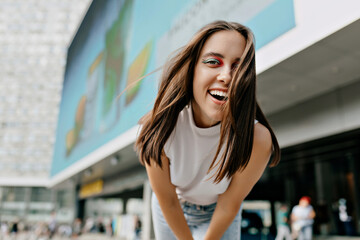 Cheerful lady with bright fashion make up ,posing at camera close up and smiling on city background. Her hair is flying in wind,