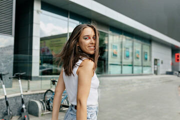 Portrait from back of stylish pretty woman with dark hair and bright make up looking at camera and walking in the city