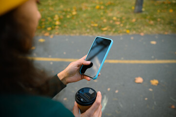 Focus on mobile phone with black empty blank screen with copy space for advertisement in the hands of blurred mature woman holding takeaway paper cup and enjoying autumn weather resting in forest park