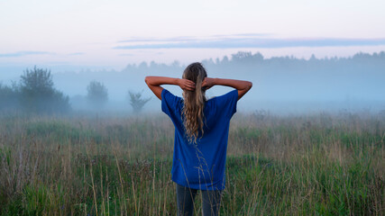 A girlin a blue t-shirt walks through a field with fog at sunset. Fog or mist over the field. Misty evening scene. Twilight fog.