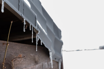 Icicles hanging from a sheet metal roof, brown blurred boards in the background. Winter weather and freezing temperatures.