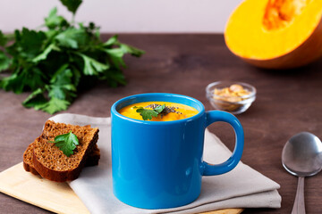Pumpkin cream soup with greens in a blue mug with spoon on the dark wooden background. soup-puree with cream, croutons and pieces of ripe pumpkin on the cutting board. copy space.