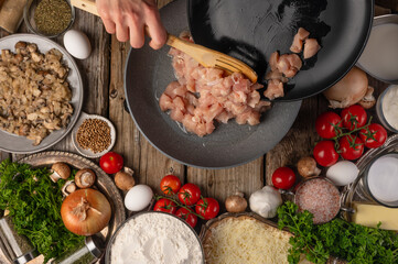 The cook fries minced chicken meat in a skillet. Lots of ingredients for making pizza, pie, ravioli. Italian cuisine, culinary blog, instagram, cookbook. High angle view.