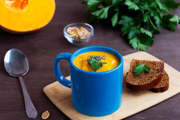 Pumpkin cream soup with greens in a blue mug with spoon on the dark wooden background. soup-puree with cream, croutons and pieces of ripe pumpkin on the cutting board. copy space