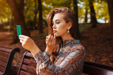 Portrait of woman applying lipstick using hand mirror in fall park sitting on bench. Girl checking makeup outdoors