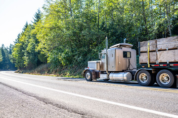 Classic big rig bonnet semi truck with roof spoiler transporting harvest in wood boxes fastened on flat bed semi trailer running for delivery on the highway road with trees on the hill