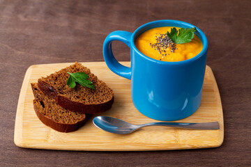 Pumpkin cream soup with greens in a blue mug with bread on the dark wooden background. vegetarian soup-puree with cream, croutons and spoon on the cutting board.