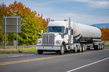 Classic industrial white semi truck transporting liquefied gas in a specialized high-pressure tank semi trailer driving on the road with yellow and red autumn trees