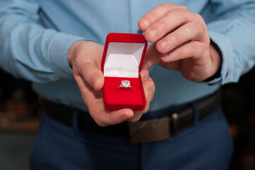 An open red velvet box with a white gold ring with a precious stone in the hands of a man in a blue shirt
