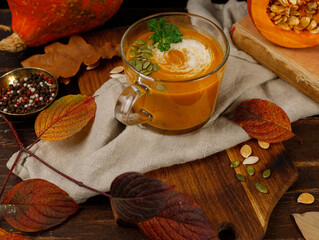 Vegetable Pumpkin cream soup in a glass mug. Close-up on a dark wooden background, with bright autumn leaves, horizontal