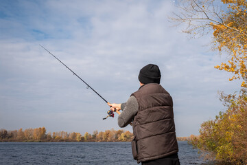 A fisherman with a spinning rod on the bank of the river is fishing. A man with a fishing rod in nature, view from the back