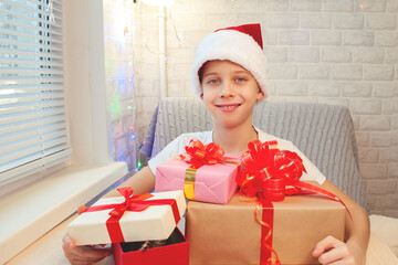 A child in a Santa Claus hat laughs and hugs boxes with Christmas gifts. Made by hand. Bright interior. Home leisure with children in the New Year.
