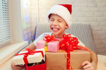 A child in a Santa Claus hat laughs, makes faces and hugs boxes with Christmas gifts. Made by hand. Bright interior. Home leisure with children in the New Year.