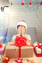 A child in a Santa Claus hat has closed his eyes with his hands and is waiting for a New Year's gift. A large box with a red bow stands in front of the boy.