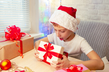 A child in a Santa Claus hat looks in surprise into an open gift box. Christmas interior of the house.