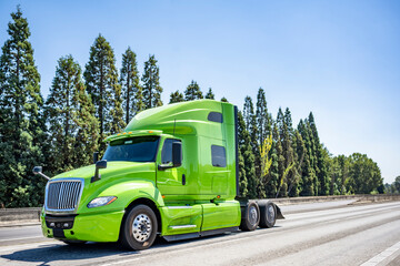 Fototapeta na wymiar Light green big rig professional semi truck tractor driving on the wide multiline highway road to warehouse for pick up loaded semi trailer for the next freight