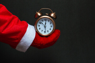 Festive concept. In the hand of santa claus an alarm clock of copper color on a black background. close-up