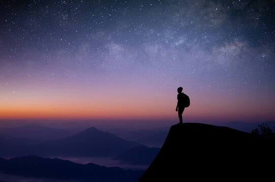 Silhouette of young traveler with backpack standing alone on top of the mountain and watched the beautiful view night sky, star and milky way. He was happy to travel alone in the wide world.