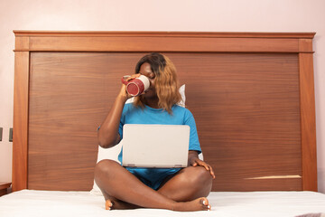 young african woman using her laptop in bed, drinking from a coffee mug