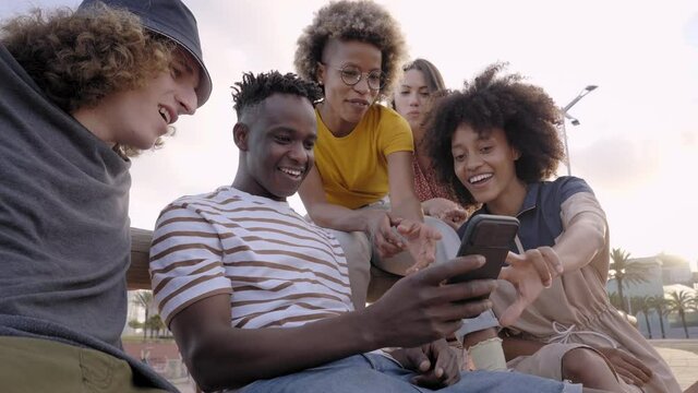 Happy smiling teenage friends laughing outside at something in smartphone or mobile phone. Young multiracial people spending time together. Friendship, communication, youth and lifestyle concept