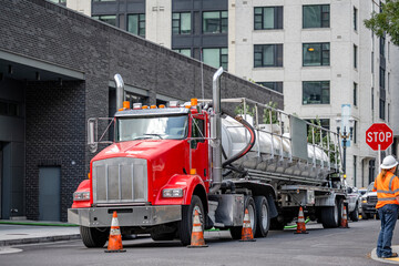 Red classic big rig semi truck with utility tank semi trailer standing on the city street with road...