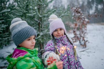 Happy kids with burning sparklers during walking in forest on snowing day, outdoor family Christmas celebration, excited family in warm clothes