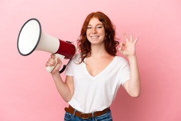 Teenager reddish woman isolated on pink background holding a megaphone and showing ok sign with fingers