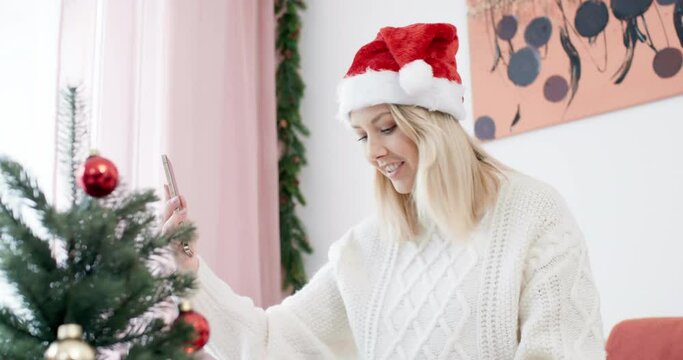 Young smiling woman wearing a santa hat making video call on cosy couch and opens christmas gift in front of selfi cam shot in 4k