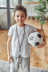 Full length portrait of smiling kid wearing referee whistle and holding ball for match at home