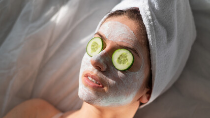 A woman with a towel on her hair and in a clay face mask and cucumbers in front of her eyes lies on...