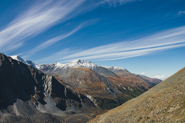 Colorful alpine landscape with great mountain in autumn colors with snow on peak in sunshine under cirrus clouds in blue sky. Picturesque autumn scenery with sunlit rocks and snow-covered mountain top