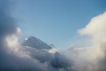 Beautiful view of snow-capped mountains above thick clouds in sunshine. Scenic bright mountain landscape with white-snow peak among dense low clouds in blue sky. Wonderful scenery with snowy pinnacle.