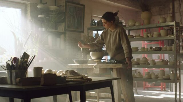 Woman potter is standing near pottery wheel in art studio, removing excessive water from raw clay pot with a sponge, dries the item before putting it to a kiln, ceramic art, Slow motion.