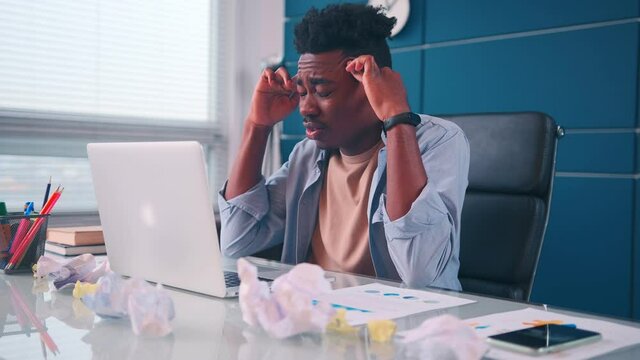 Stressed young African American businessman sitting at table with laptop full of crumpled paper documents, suffering from lack of motivation or inspiration, stack with difficult task.
