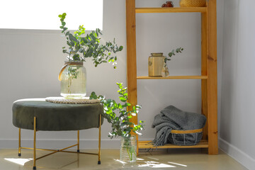 Vases with green eucalyptus branches on pouf near light wall
