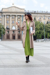 Young woman in leather cap holding newspaper and walking on city square