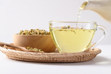 Chamomile tea in a teapot pouring into cup glass with dried chamomile flowers in a bowl on white...
