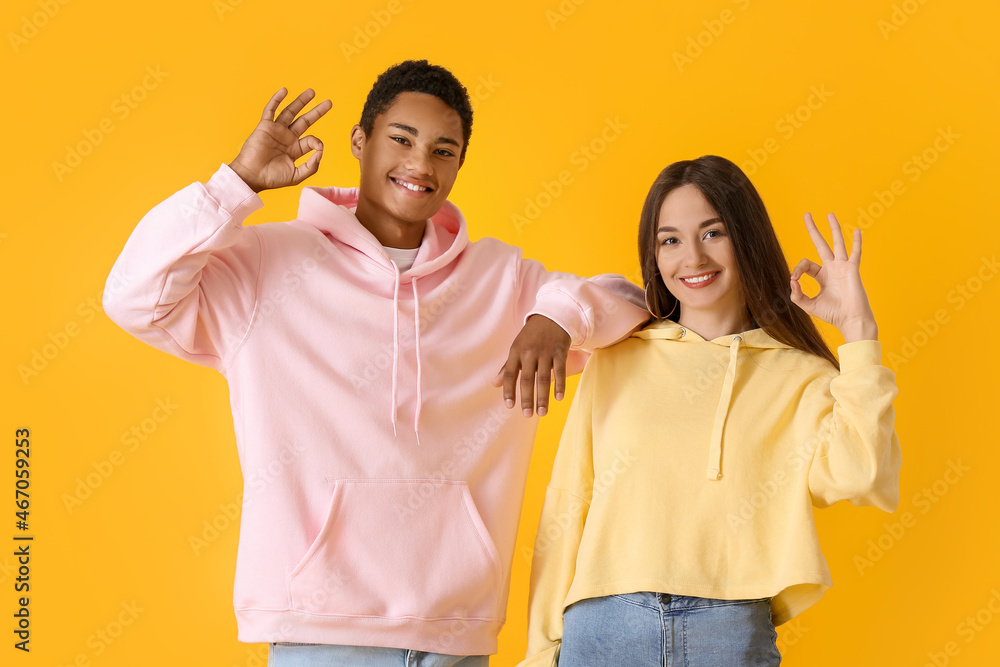 Wall mural Stylish young couple in hoodies showing OK on yellow background - Wall murals