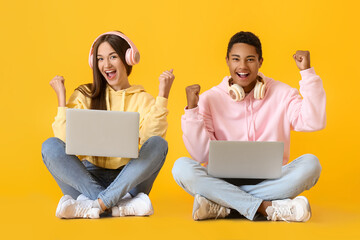 Stylish young couple in hoodies using laptops on yellow background