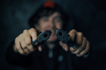 Two pistols in man's hands are pointed at camera. Guy threatens with firearm. Criminal with weapon. Murderer in hood on dark background. Close-up of two gun muzzles.