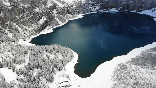 Aerial flyover next to the shores of lake Oeschinensee in Kandersteg, Switzerland on a winter day with mountain peaks hidden clouds