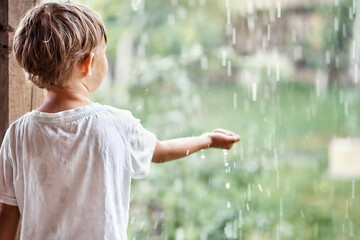 Blond little boy in white t-shirt holds hand under falling down rain drops standing on country house terrace backside view